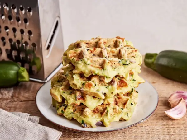 Stapel goldbrauner Zucchini-Waffeln auf einem Teller, daneben Zucchini, Knoblauch und eine Reibe.
