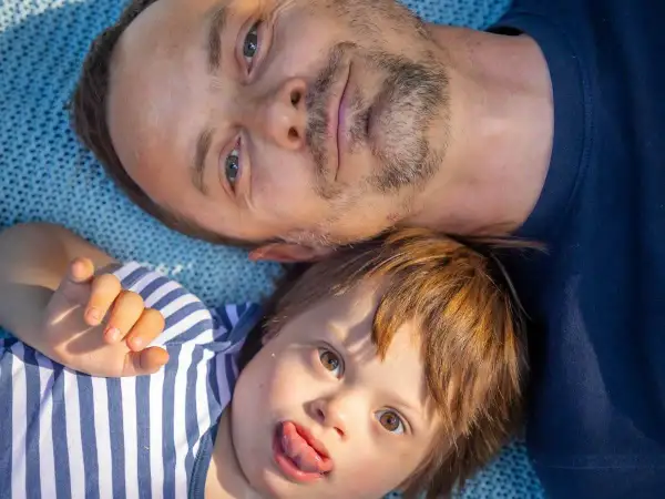 Un padre e il suo bambino con sindrome di Down, un neonato con un'espressione giocosa, sdraiati su una coperta blu.