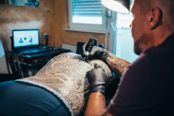 Artysta tworzy skomplikowany tatuaż na plecach klienta. Widać detale wzoru i ręce tatuażysty w czarnych rękawiczkach.