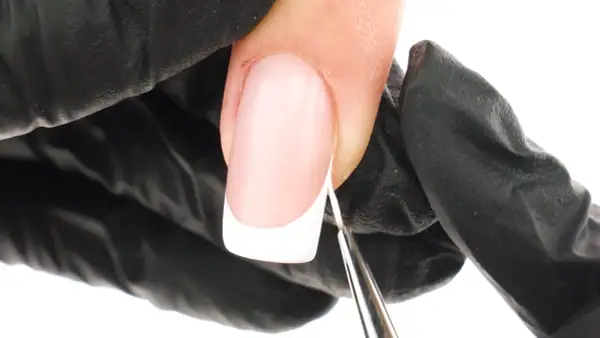 Precyzyjne malowanie białej końcówki french manicure bez użycia pasków. Dłoń w czarnej rękawiczce trzyma pędzelek.