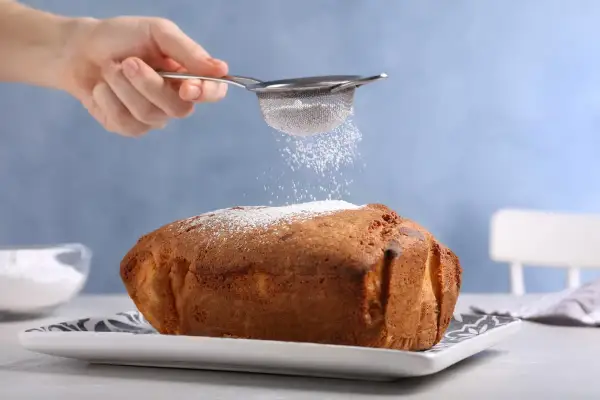 Una mano setaccia zucchero a velo su un dolce appena sfornato, un tocco finale perfetto per ogni ricetta Bimby.