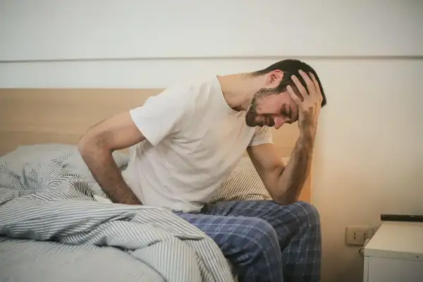 Mann sitzt im Bett, hält sich den Kopf. Mögliche Bewegungsmangel Symptome wie Kopfschmerzen und Müdigkeit sind zu erkennen.