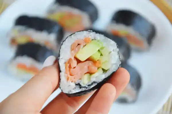 Smaczny futomak z łososiem i awokado, trzymany w dłoni na talerzu pełnym sushi.