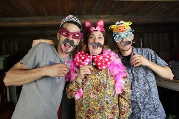 Tres amigos se divierten con disfraces hechos con cosas de casa: un hombre con tiara y bigote, una mujer con orejas de gato y un hombre con máscara de vaca.