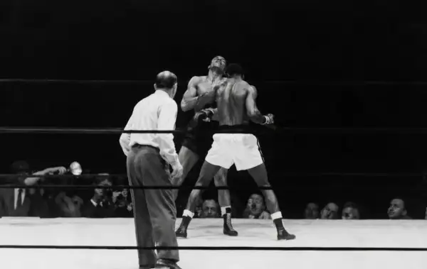 A referee watches as two boxers battle in the ring. One boxer, with his head thrown back, is hit. This intense moment captures the fight for who is the king of boxing.