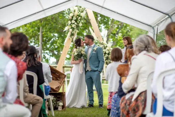 Un couple s'embrasse lors de leur baptême civil, entouré de leurs proches sous une tente décorée de fleurs.