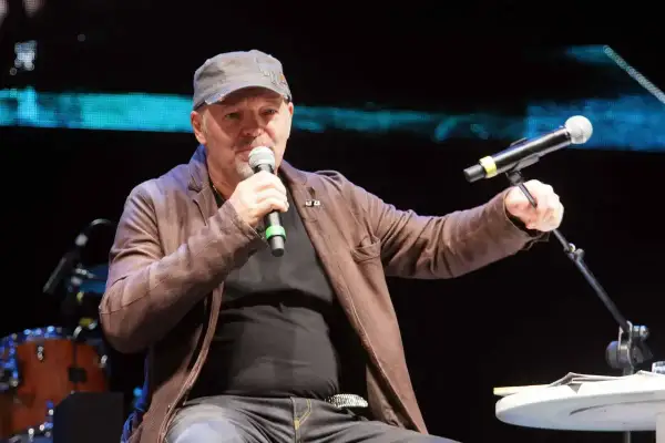 Uomo con cappellino e giacca di pelle marrone parla al microfono, sembra che sono ancora in coma, ma è sul palco.