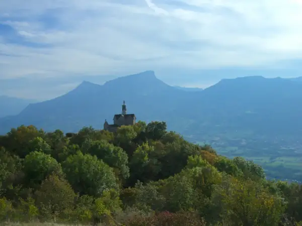Un petit Mont Saint Michel savoyard, une chapelle perchée sur une colline boisée, domine un paysage de montagnes bleutées sous un ciel nuageux.