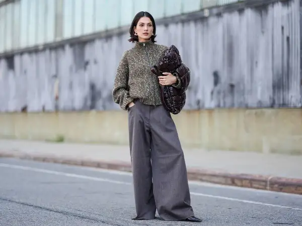 Stylisches Outfit: Eine Frau kombiniert eine graue Hose mit einem gemusterten Strickpullover und einer voluminösen Tasche.