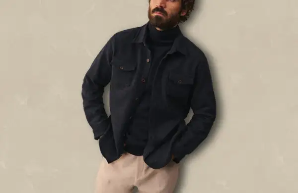 Un homme porte une surchemise foncée ouverte sur un col roulé bleu marine, avec un pantalon beige. Idéal pour savoir comment porter une surchemise avec style.