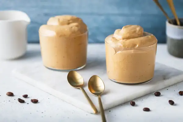 Due bicchieri di cremosa schiuma di caffè, perfetti per una ricetta crema di caffè senza panna. Due cucchiaini dorati e chicchi di caffè completano la scena.