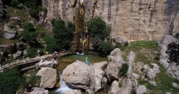 Un hermoso salto de agua cae en una poza natural rodeada de rocas y vegetación. Varias personas disfrutan del paisaje y del agua.