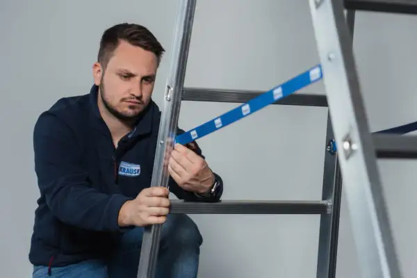 Mężczyzna w granatowej bluzie z logo KRAUSE przeprowadza badanie drabin, sprawdzając mocowanie niebieskiej taśmy zabezpieczającej.