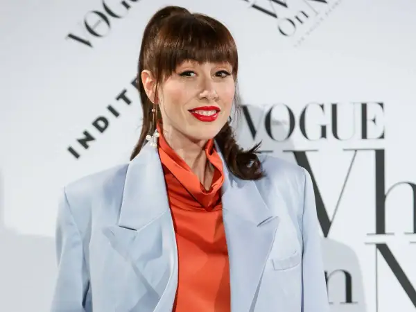 Natalia Ferviu, con su coleta alta y labios rojos, luce un traje azul pálido y un top naranja vibrante.