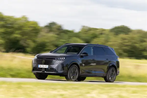 Ciemnoszary Peugeot 5008 pędzi po drodze, otoczony zielenią pól i drzew.