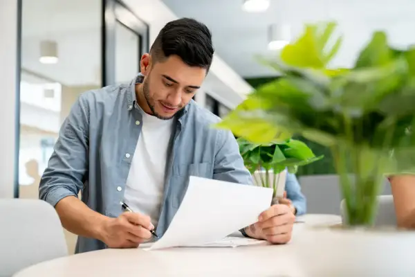 Giovane uomo concentrato su documenti, con una pianta rigogliosa che suggerisce un ambiente di lavoro moderno e un uso foresteria.