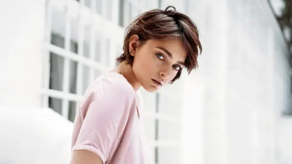 Junge Frau mit einem lässigen Pixie-Cut-Haarschnitt, blickt nachdenklich in die Kamera. Sie trägt ein zartrosa Oberteil.