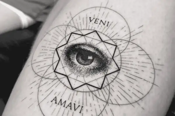 Ein filigranes Fineline Tattoo am Oberarm einer Frau zeigt ein Auge inmitten geometrischer Formen und der Worte "VENI AMAVI".