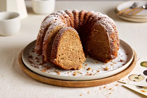 Ein saftiger Kuchen mit Dinkelmehl, bestäubt mit Puderzucker, auf einem Teller mit Holzrand. Ein Stück ist herausgeschnitten.