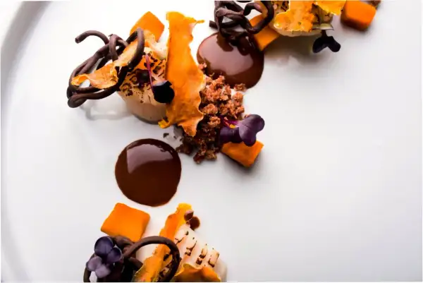 Postre de autor con chocolate, naranja y flores moradas. Una obra de arte culinaria que promete una experiencia gastronómica inolvidable.