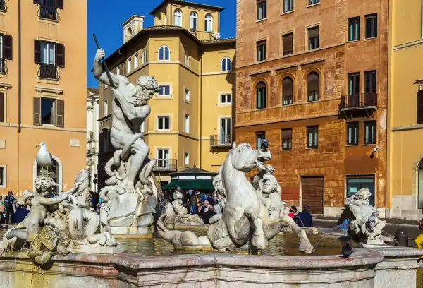 La Fuente de Neptuno en Roma, con su imponente estatua de mármol luchando contra un pulpo, rodeada de caballos marinos y figuras mitológicas, bajo el sol y edificios coloridos.