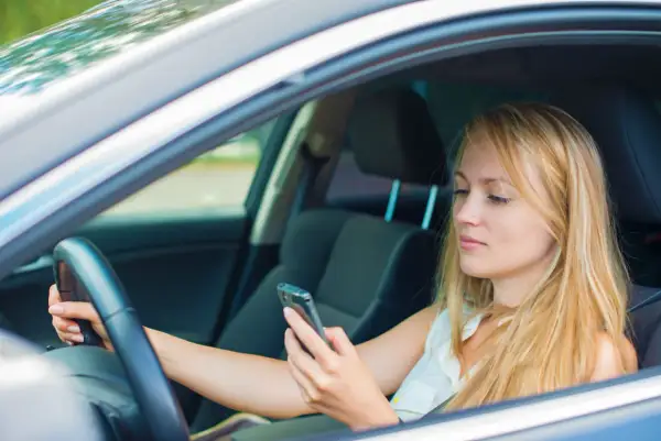 Mujer rubia al volante, mirando su móvil. Al distraerse el conductor tarda, poniendo en riesgo su seguridad y la de otros.
