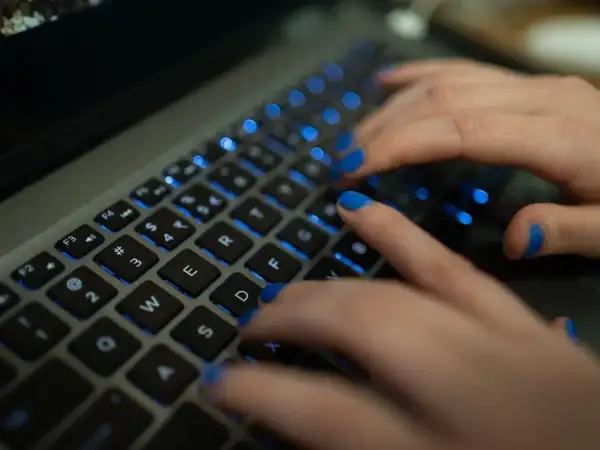 Dłonie z niebieskim lakierem na paznokciach piszą na podświetlanej klawiaturze MacBooka. Twarda spacja jest widoczna.