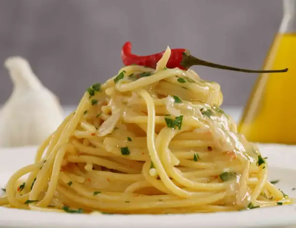 Spaghetti aglio olio e peperoncino cremosi, guarniti con un peperoncino rosso e prezzemolo. Sullo sfondo, aglio e olio d'oliva.