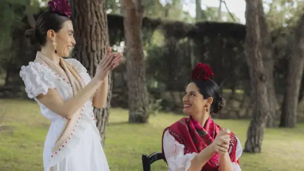 Dos mujeres vestidas para ir al Rocío, con trajes blancos y mantones, sonríen y aplauden en un jardín.