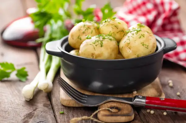 Młode ziemniaki w czarnej misce, posypane koperkiem, obok widelca i dymki.