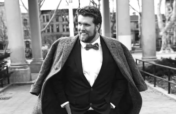 Un homme gros bien habillé, souriant, porte un costume sombre, une chemise blanche et un nœud papillon. Son manteau est jeté sur ses épaules.