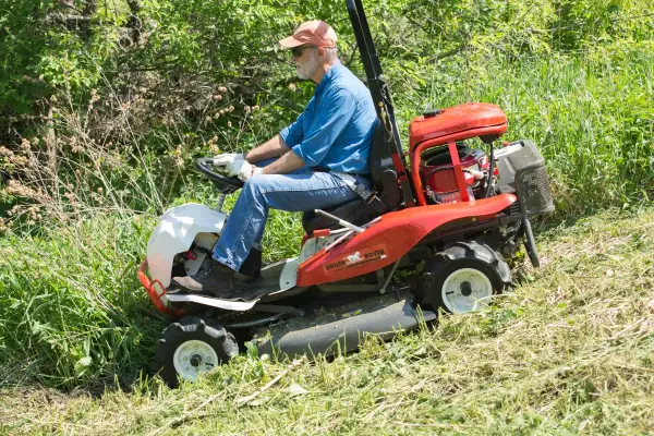 Mężczyzna w czapce i okularach prowadzi kosiarkę Brush Rover, kosząc wysokie trawy. Dobre opinie o dedra sprzęcie potwierdzają jego skuteczność.