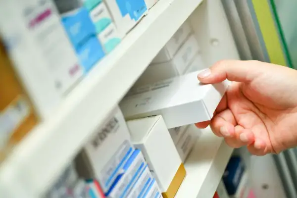 Dłoń sięga po białe pudełko leku, być może zawierającego nilogrin, z aptecznej półki pełnej innych opakowań.
