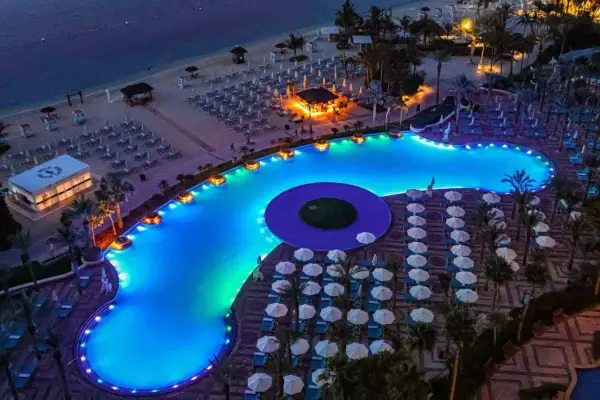 Nocny widok na luksusowy basen i plażę w Dubaju. Błękitna woda basenu, oświetlona kolorowymi światłami, otoczona leżakami i palmami.