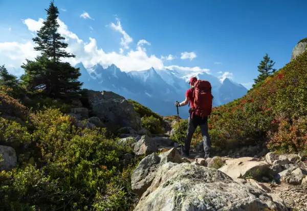 Randonneur avec sac à dos rouge, bâtons de marche, sur un sentier rocheux. Montagnes enneigées en arrière-plan. Combien de jours de marche pour atteindre ce sommet ? gr5.