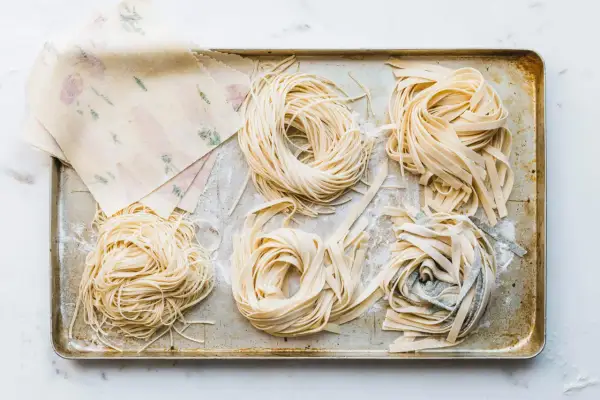 Frische Nudeln in verschiedenen Formen liegen auf einem Blech. Ein Stapel mit Blumenmuster verzierter Teigblätter liegt daneben. Perfekt für alle, die Nudeln selber machen wollen.