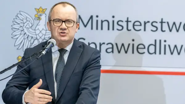 Mężczyzna w garniturze przemawia przy mikrofonie na tle godła Polski i napisu "Ministerstwo Sprawiedliwości". Może mówić o problemach z mieszkaniem komunalnym w Warszawie.