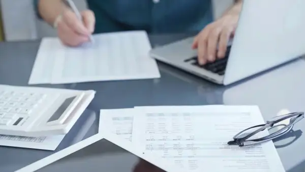 Osoba pracuje przy laptopie, przeglądając dokumenty i pisząc. Na biurku leży kalkulator i okulary. Przygotowania do podpisania umowy zlecenie 2026.