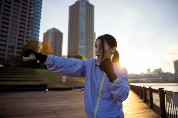 A woman practices shadow boxing outdoors, a popular fitness trend, with city buildings in the background at sunset.