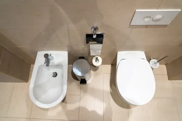 Łazienka z bidetem i WC. Widać kosz na śmieci, papier toaletowy i szczotkę. Odległość WC od ściany bocznej jest optymalna.