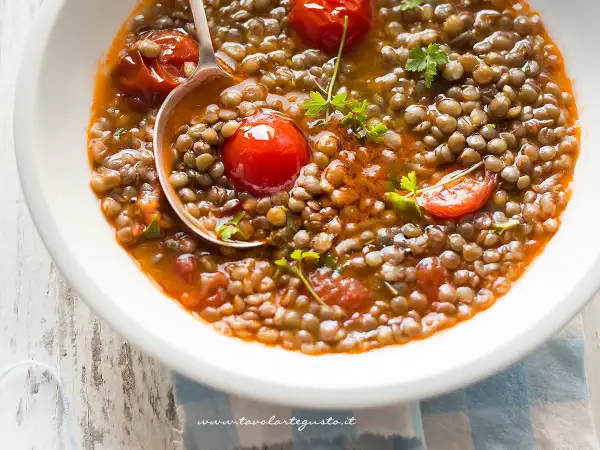 Una ciotola bianca piena di saporita zuppa di lenticchie con pomodorini e prezzemolo fresco. Un mestolo serve la zuppa.