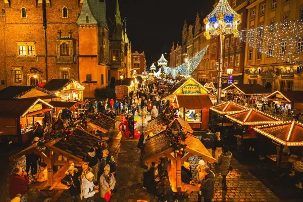 Tłoczny wrocławski jarmark bożonarodzeniowy nocą, rozświetlony tysiącami lampek. Ludzie spacerują między drewnianymi straganami, ciesząc się świąteczną atmosferą.