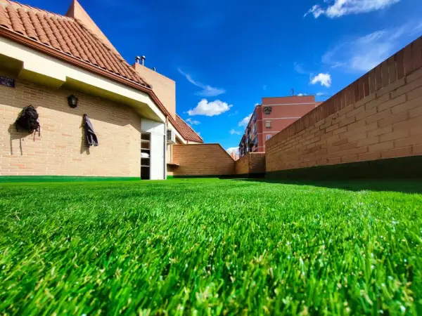 Un vibrante césped artificial para parques infantiles cubre una terraza con edificios de ladrillo y un cielo azul brillante.