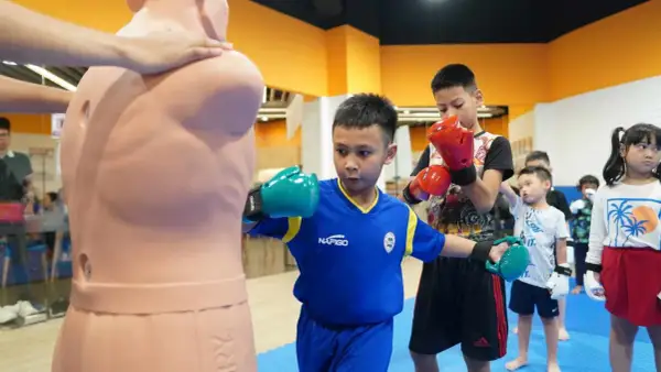 Young boxers practice their moves, with one boy demonstrating boxing blocking techniques on a training dummy.