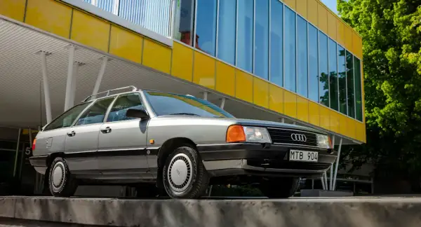 En grå Audi 100 C3 Avant står parkerad framför en modern gul byggnad med stora fönster.