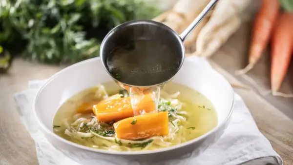 Un mestolo versa brodo caldo in una ciotola di zuppa di pollo con carote e tagliatelle.