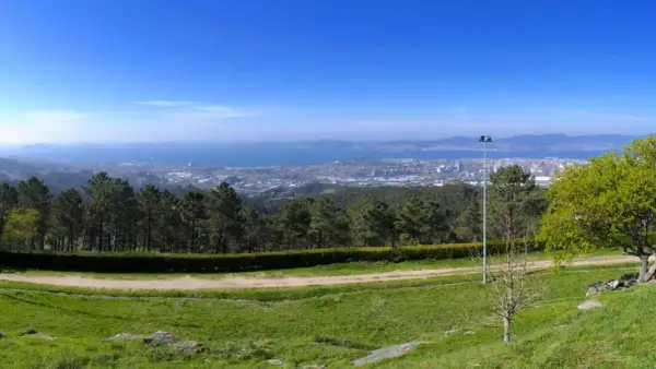 Vista panorámica desde el monte dos pozos, con un camino de tierra y césped en primer plano, árboles y un faro, y una ciudad y el mar al fondo.