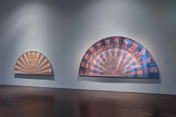 Two fan-shaped artworks display intricate pattern and decoration, creating a sense of movement. The larger piece bursts with colorful fabric squares, while the smaller one features a softer, sunburst design.