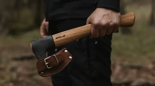 Mano sujetando hachas tácticas con funda de cuero. El mango de madera tiene una inscripción.