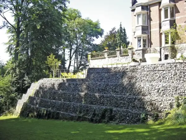 Imponująca skarpa na granicy działki, zbudowana z kamieni w gabionach, tworzy tarasy porośnięte zielenią. Na górze widać fragment rezydencji.
